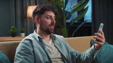 Abur cubur reklamı konsepti. Sakallı adam evde fast food dağıtımı ile vakit geçiriyor, kanepede oturuyor, akıllı telefondan video konuşmaları yapıyor ve kola içiyor..