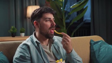 Abur cubur reklamı konsepti. Sakallı adam evde fast food dağıtımı ile vakit geçiriyor, kanepede oturup TV 'de film izliyor, gülümsüyor ve soslu kızarmış patates yiyor..