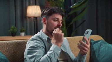 Abur cubur reklamı konsepti. Sakallı adam evde fast food dağıtımı ile vakit geçiriyor, kanepede oturup akıllı telefondan spor maçı izliyor ve cips yiyor, hayal kırıklığına uğramış yüz ifadesi..