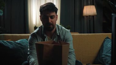 Abur cubur reklamı konsepti. Yetişkin sakallı bir adam evde vakit geçirir, kanepeye oturur, elinde fast food paketiyle oturur, televizyonu açar ve uzaktan kumandayla kanalları değiştirir. Yavaş çekim.