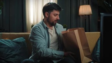 Abur cubur reklamı konsepti. Sakallı adam evde fast food teslimatlarıyla vakit geçiriyor, kanepeye oturuyor kızarmış tavuk budu ve patatesle kovayı çıkarıp masaya koyuyor. Yavaş çekim.