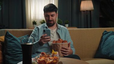 Abur cubur reklamı konsepti. Sakallı adam evde fast food teslimatlarıyla vakit geçiriyor, elinde kovayla tavuk budu tutuyor ve akıllı telefondan mesaj yazıyor. Yavaş çekim