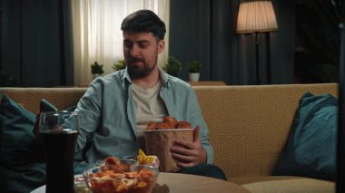 Abur cubur reklamı konsepti. Sakallı adam evde fast food teslimatlarıyla vakit geçiriyor, elinde kovayla tavuk budu tutuyor ve akıllı telefondan selfie çekiyor. Yavaş çekim