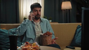 Abur cubur reklamı konsepti. Adam evde fast food teslimatçısıyla vakit geçiriyor, elinde kızarmış tavukla kanepede oturuyor ve akıllı telefondan olumsuz ifadeleri tartışıyor. Yavaş çekim