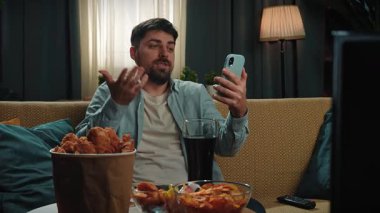 Abur cubur reklamı konsepti. Sakallı adam evde fast food dağıtımı ile vakit geçiriyor, kanepede oturmuş akıllı telefondan video konuşmaları yapıyor ve kola içiyor. Yavaş çekim
