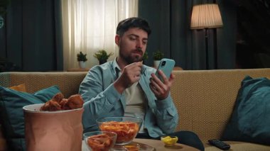 Abur cubur reklamı konsepti. Sakallı adam evde fast food dağıtımı ile vakit geçiriyor, kanepede oturup akıllı telefondan spor maçı izliyor ve cips yiyor, mutlu şarap cümleleri kuruyor. Yavaş çekim