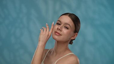 Güzellik konsepti. Makyajlı genç bir kadın stüdyoda kameraya poz veriyor, neşeli bir şekilde gülümsüyor pürüzsüz tenine dokunuyor, mavi arka planda soyutlanmış. Yavaş çekim.