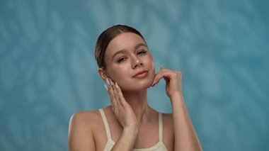 Güzellik konsepti. Makyajlı genç bir kadın stüdyoda kameraya poz veriyor, neşeli bir şekilde gülümsüyor pürüzsüz tenine dokunuyor, mavi arka planda soyutlanmış. Yavaş çekim.