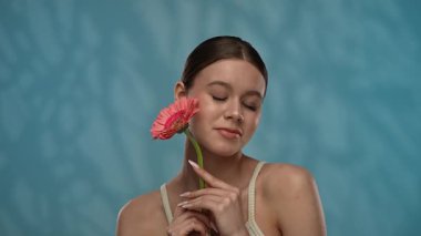 Güzellik konsepti. Makyajlı genç bir kadın stüdyoda çiçekle poz veriyor, neşeli bir şekilde gülümsüyor pürüzsüz tenine dokunuyor, mavi arka planda soyutlanıyor. Yavaş çekim.