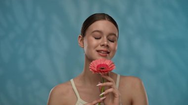 Güzellik konsepti. Makyajlı genç bir kadın stüdyoda çiçekle poz veriyor, neşeli bir şekilde gülümsüyor pürüzsüz tenine dokunuyor, mavi arka planda soyutlanıyor. Yavaş çekim.