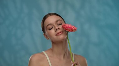 Güzellik konsepti. Makyajlı genç bir kadın stüdyoda çiçekle poz veriyor, neşeli bir şekilde gülümsüyor pürüzsüz tenine dokunuyor, mavi arka planda soyutlanıyor. Yavaş çekim.