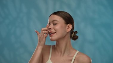 Güzellik konsepti. Makyajlı genç bir kadın stüdyoda kameraya poz veriyor, neşeli bir şekilde gülümsüyor ve burun ucuna krem sürüyor, mavi arka plana izole edilmiş. Yavaş çekim.