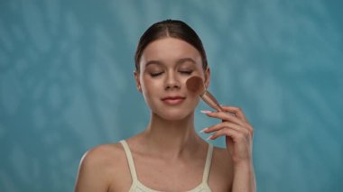 Güzellik konsepti. Makyajlı genç bir kadın stüdyoda kameraya poz veriyor, neşeli bir şekilde gülümsüyor makyaj fırçasıyla allık sürüyor, mavi arka planda izole edilmiş. Yavaş çekim.