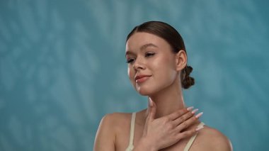 Güzellik konsepti. Makyajlı genç bir kadın stüdyoda kameraya poz veriyor, gülümsüyor ve göğüs ve boyun bölgesine krem sürüyor, mavi arka planda izole edilmiş. Yavaş çekim.