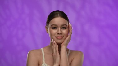 Güzellik konsepti. Stüdyoda makyajlı genç bir kadın, neşeli bir şekilde gülümsüyor pürüzsüz tenine dokunuyor, mor arka planda izole edilmiş. Yavaş çekim.