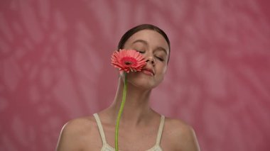Güzellik konsepti. Makyajlı genç bir kadın stüdyoda çiçekle poz veriyor, neşeli bir şekilde gülümsüyor pürüzsüz tenine dokunuyor, kırmızı arka planda izole edilmiş. Yavaş çekim.