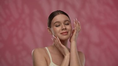 Güzellik konsepti. Makyajlı genç bir kadın stüdyoda kameraya poz veriyor, neşeli bir şekilde gülümsüyor pürüzsüz tenine dokunuyor, kırmızı arka planda izole edilmiş. Yavaş çekim.