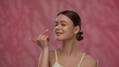 Güzellik konsepti. Makyajlı genç bir kadın stüdyoda kameraya poz veriyor, neşeli bir şekilde gülümsüyor ve burun ucuna krem sürüyor, kırmızı arka planda izole edilmiş. Yavaş çekim.