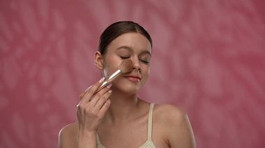 Güzellik konsepti. Makyajlı genç bir kadın stüdyoda kameraya poz veriyor, neşeli bir şekilde gülümsüyor makyaj fırçasıyla allık sürüyor, kırmızı arka planda izole edilmiş. Yavaş çekim.