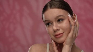 Güzellik konsepti. Makyajlı genç bir kadın stüdyoda kameraya poz veriyor, neşeli bir şekilde gülümsüyor pürüzsüz tenine dokunuyor, kırmızı arka planda izole edilmiş bir şekilde kapanıyor. Yavaş çekim.