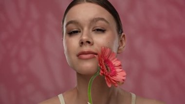 Güzellik konsepti. Makyajlı genç bir kadın stüdyoda çiçekle poz veriyor, neşeli bir şekilde gülümsüyor pürüzsüz tenine dokunuyor, kırmızı arka planda izole edilmiş. Yavaş çekim.
