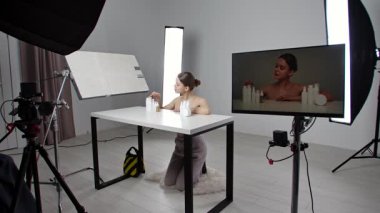 Stüdyoda profesyonel kozmetik klip çekiminin kulisinde. Genç bir kız beyaz bir stüdyoda kozmetik ürünleriyle kavanozları gösteriyor. Kameraman kamerayı açar, aydınlatma ekipmanları görünür..