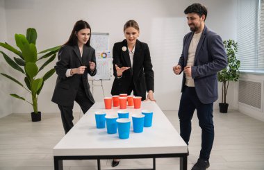 Bir grup iş arkadaşı ofis ortamında fincanlarla eğlenceli bir oyun oynuyor, takım çalışmasını ve işbirliğini teşvik ediyor.