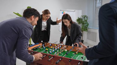Bir grup ofis çalışanı parlak ve modern bir çalışma alanında yoğun bir şekilde masa futbolu oynuyorlar. İfadeleri heyecan ve odaklanma gösteriyor..