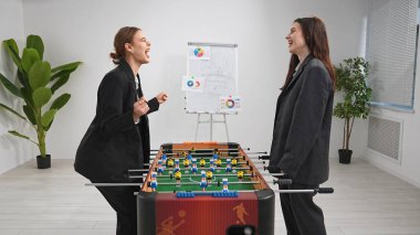 Takım elbiseli iki kadın ofiste langırt oynamaktan hoşlanıyor. Ortam canlı ve rekabetçi, takım çalışmalarını ve dostluklarını vurguluyorlar..