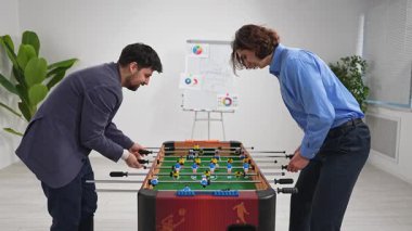 Modern bir ofis alanında langırt maçına katılan iki iş arkadaşı, takım çalışması ve rekabet sergiliyor..