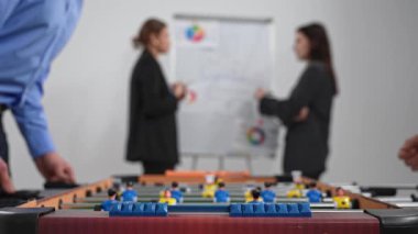 Ön planda bir langırt masası görülebilirken, iki iş adamı bir tablonun yanında tartışıyorlar. Kapat.