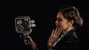Stüdyoda profesyonel model. Siyah arka planda izole edilmiş kameraya poz veren eski moda saç stili ve makyajlı kadın oyuncu eski video kamerada gülümseyen ve selfie çeken kadın..
