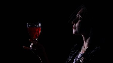 Stüdyoda profesyonel model. Siyah arka planda poz veren eski moda elbiseli kadın oyuncu silueti, kırmızı şarap kadehiyle peçeli kadın..