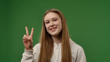 Gençlik duyguları konsepti. Kameraya poz veren genç kız zafer işareti ve mutluluk işareti gösteriyor, olumlu ifade kazanıyor, krom anahtar yeşil ekranlı arka plan modelinde izole edilmiş. Yavaş çekim