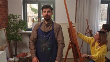 Sanatsal beceriler geliştirmek için eğitim boş zamanı aktivitesi. Stüdyoda yüksek lisans sırasında resim yapan bir grup insanın önünde önlüklü resim öğretmeni pozu, olumlu ifade. Yavaş çekim