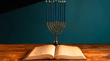 Güzel aydınlatılmış bir menorah ve kırsal bir ahşap masaya karşı açık bir kitap. Sıcak parıltı, Hanuka bayramında gelenek ve inancı simgeliyor..