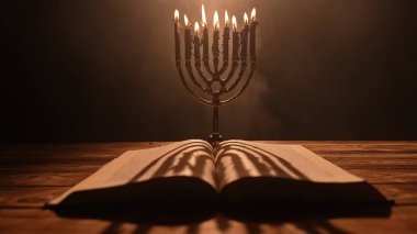 Çok güzel aydınlatılmış bir menorah, titreyen mum ışığının ortasında, kırsal bir ahşap masaya yerleştirilmiş açık bir kitaba gölgeler seriyor..