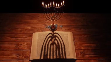 Güzel aydınlatılmış bir menorah gururla eski bir ahşap masanın üzerinde durur, antik bir metnin üzerine karmaşık gölgeler bırakır..