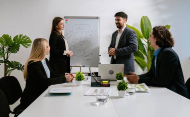 Profesyonel bir iş ekibi modern bir ofiste stratejiyi tartışır. Takım elbiseli bir adam, tahtada fikirleri özetliyor ve olumlu sonuç için bayan meslektaşına teşekkür ediyor. Meslektaşlar başarıya seviniyor..