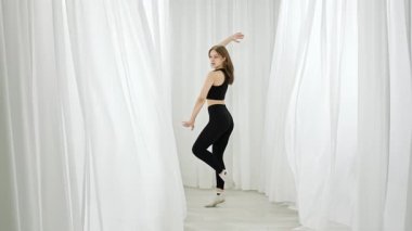 Siyah elbiseli bir dansçı zarif bir şekilde, parlak, minimalist beyaz bir stüdyo odasında, zarafet göstererek performans sergiler. Yavaş çekim.