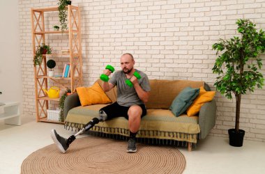 Özürlü insanların günlük hayatı ve günlük rutini. Biyonik protez bacak sakatlığı olan bir adam kanepede oturup dambıllarla spor egzersizi yapıyor..