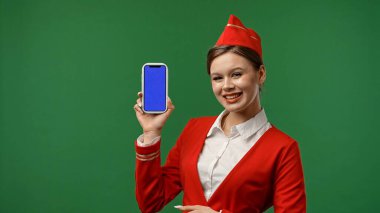 Uçuş ekibi servis ilanı. Hostes gibi giyinmiş kırmızı üniformalı bir kadın elinde mavi ekranlı akıllı telefon, krom anahtar yeşil ekranlı stüdyo arka planında izole edilmiş..