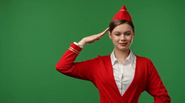 Uçuş ekibi reklam konsepti. Hostes gibi giyinmiş bir kadın kırmızı üniformalı kameraya selam veriyor, olumlu ifade, krom anahtar yeşil ekran stüdyo arka planında izole edilmiş..