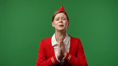 Uçuş ekibi reklam konsepti. Hostes kılığındaki kırmızı üniformalı kadın lütfen diyor ve dua jesti ile el ele tutuşuyor. Renkli yeşil ekran stüdyo arka planında izole edilmiş..