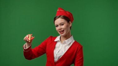 Uçuş ekibi reklam konsepti. Kırmızı üniformalı hostes gibi giyinmiş bir kadın oyuncak uçakla oynuyor ve bunu gösteriyor. Renkli yeşil ekranlı stüdyo arka planında izole edilmiş..