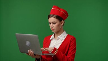 Uçuş ekibi reklam konsepti. Kırmızı üniformalı hostes kılığındaki bir kadın dizüstü bilgisayarını tutuyor ve klavyede daktilo kullanıyor. Krom anahtar yeşil ekranlı stüdyo arka planında izole edilmiş..