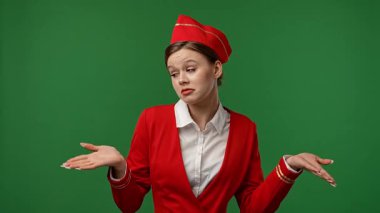 Uçuş ekibi reklam konsepti. Hostes gibi giyinmiş kırmızı üniformalı bir kadın. İfadesiz bir ifade ve omuz silkerek poz veriyor. Renkli yeşil ekranlı stüdyo arka planında izole edilmiş..