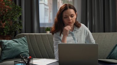 Günlük yaşam tarzı konsepti. Genç, yetişkin bir iş kadını, oturma odasında oturmuş dizüstü bilgisayarla çalışıyor, ekrana bakıyor..