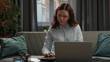 Günlük yaşam tarzı konsepti. Genç yetişkin iş kadını ev ofisinde vakit geçiriyor, oturma odasında oturan kadın dizüstü bilgisayarla çalışıyor, hesaplamalar yapıyor..