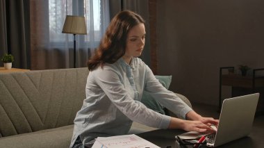 Günlük yaşam tarzı konsepti. Genç yetişkin iş kadını ev ofisinde vakit geçiriyor. Oturma odasında oturan, dizüstü bilgisayarda çalışan, klavyede yazan bir kadın..